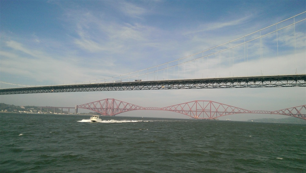 A speedboat passes under the Forth road bridge with the iconic red rail bridge in the distance